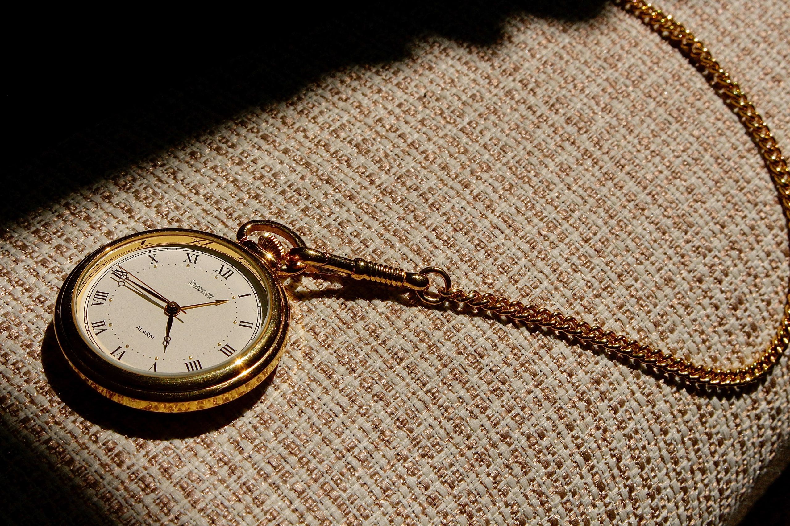 A vintage gold pocket watch with a chain resting on woven fabric, bathed in natural sunlight.